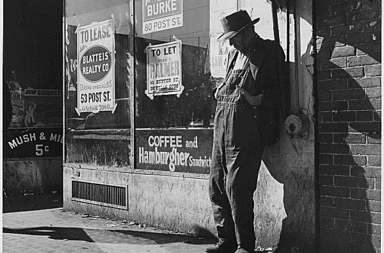 Great Depression: unemployed, destitute man leaning against vacant store, San Francisco (1935). By Dorothea Lange. Via Wikimedia Commons.