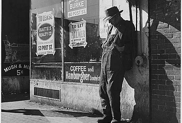 Great Depression: unemployed, destitute man leaning against vacant store, San Francisco (1935). By Dorothea Lange. Via Wikimedia Commons.