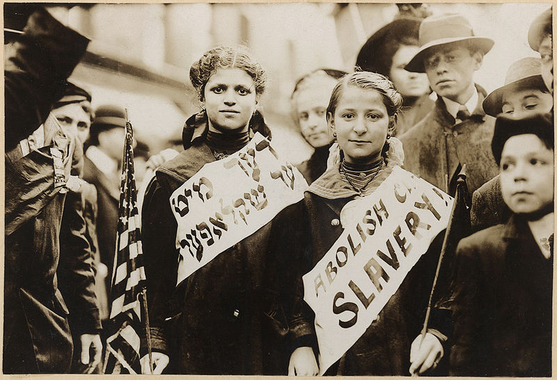 Two girls wearing banners with slogan "ABOLISH CH[ILD] SLAVERY!!" in English and Yiddish. one carrying American flag; spectators stand nearby. Probably taken during May 1, 1909 labor parade in New York City.