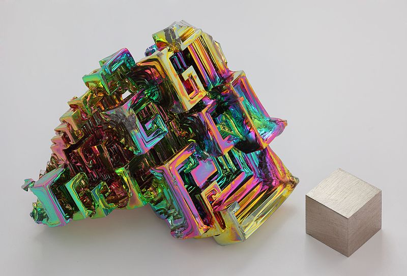 The chemical element bismuth as a synthetic made crystal. The iridescent surface is a very thin layer of oxidation. Beside it is a high purity (99.99 %) 1 cm3 bismuth cube for comparison. By Alchemist-hp