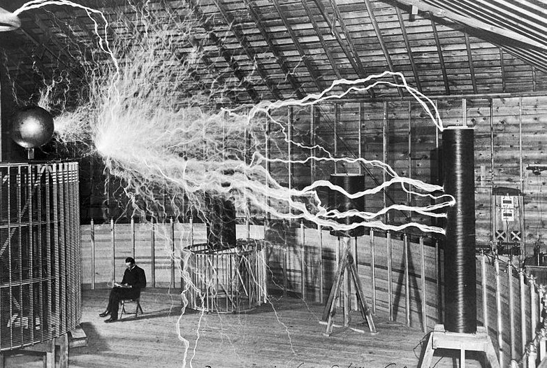 Nikola Tesla sitting in his Colorado Springs laboratory next to his huge "magnifying transmitter" Tesla coil which is producing 22 foot bolts of electricity