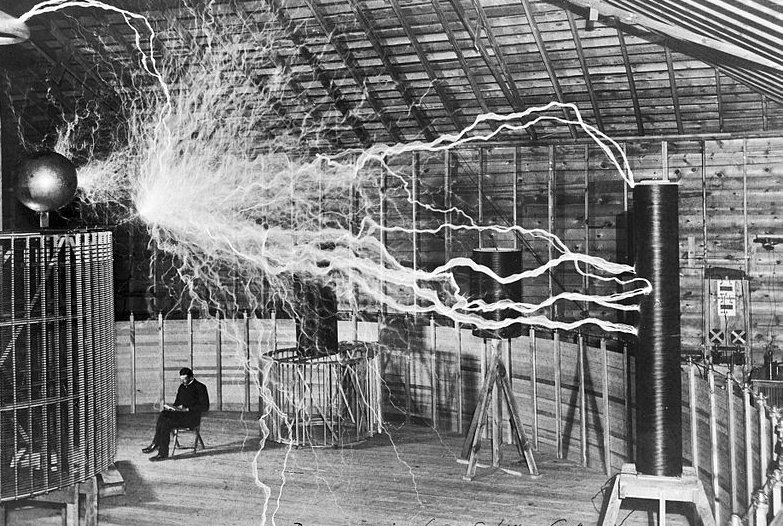 Nikola Tesla sitting in his Colorado Springs laboratory next to his huge "magnifying transmitter" Tesla coil which is producing 22 foot bolts of electricity