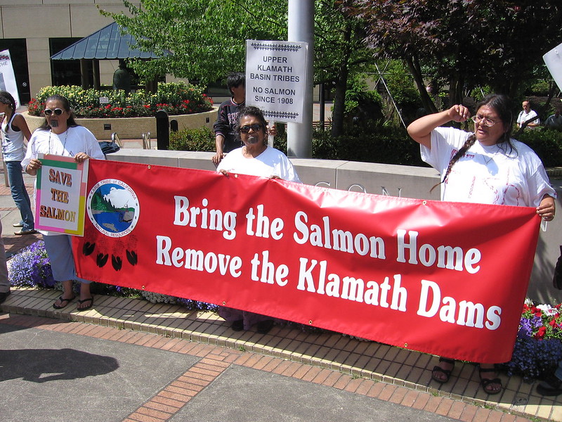 Three women hold sign saying "bring the salmon home, remove the Klamath Dams."